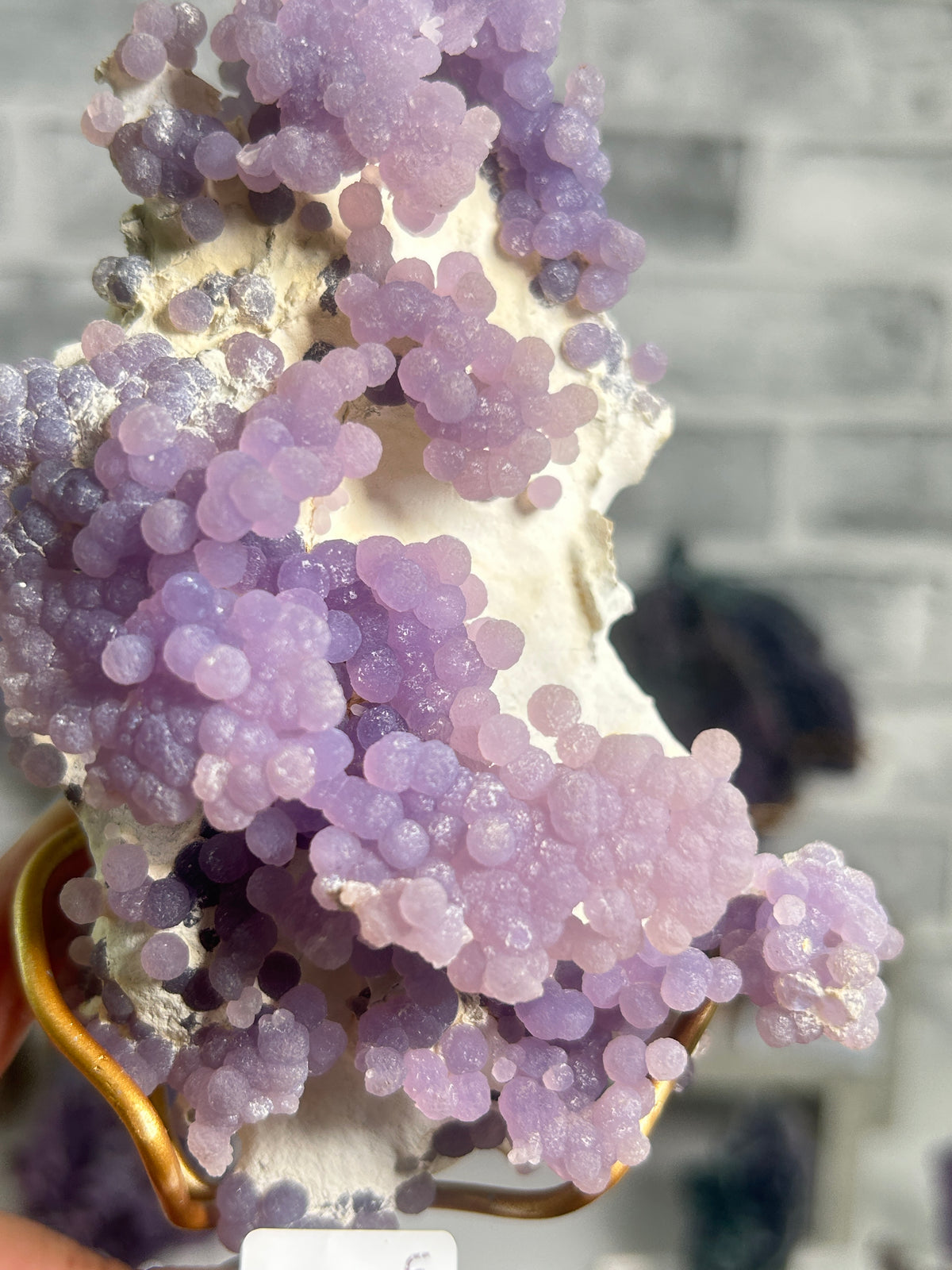 A close-up of a lavender and lilac crystal cluster with rounded, bubble-like formations on a white matrix, held in a gold-colored ring holder against a blurred gray brick wall background.