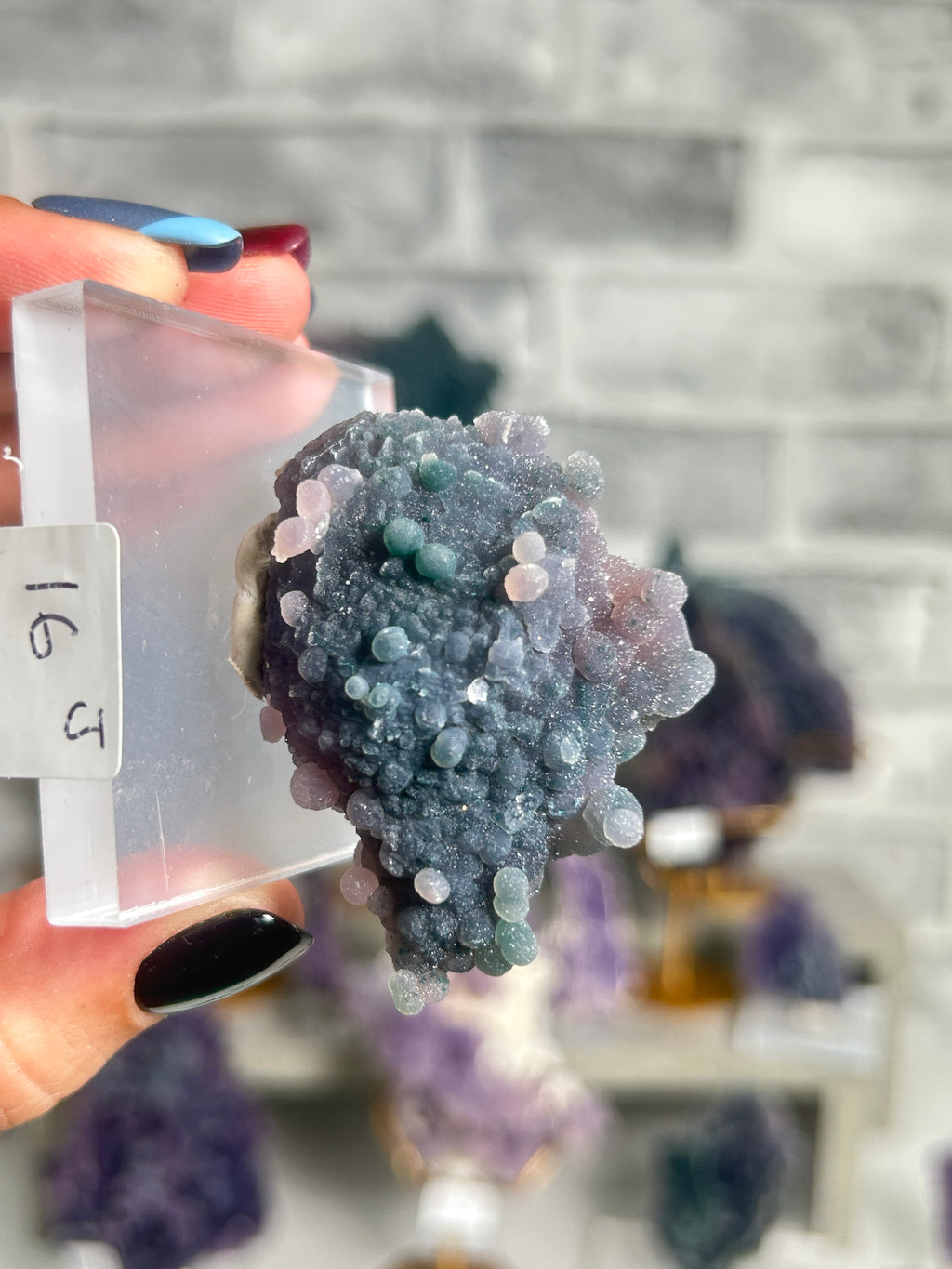 A hand with dark and blue nail polish holds a clear acrylic block displaying a textured geode specimen with blue, purple, and pink crystalline formations. The background is a blurred gray brick wall with other mineral specimens.
