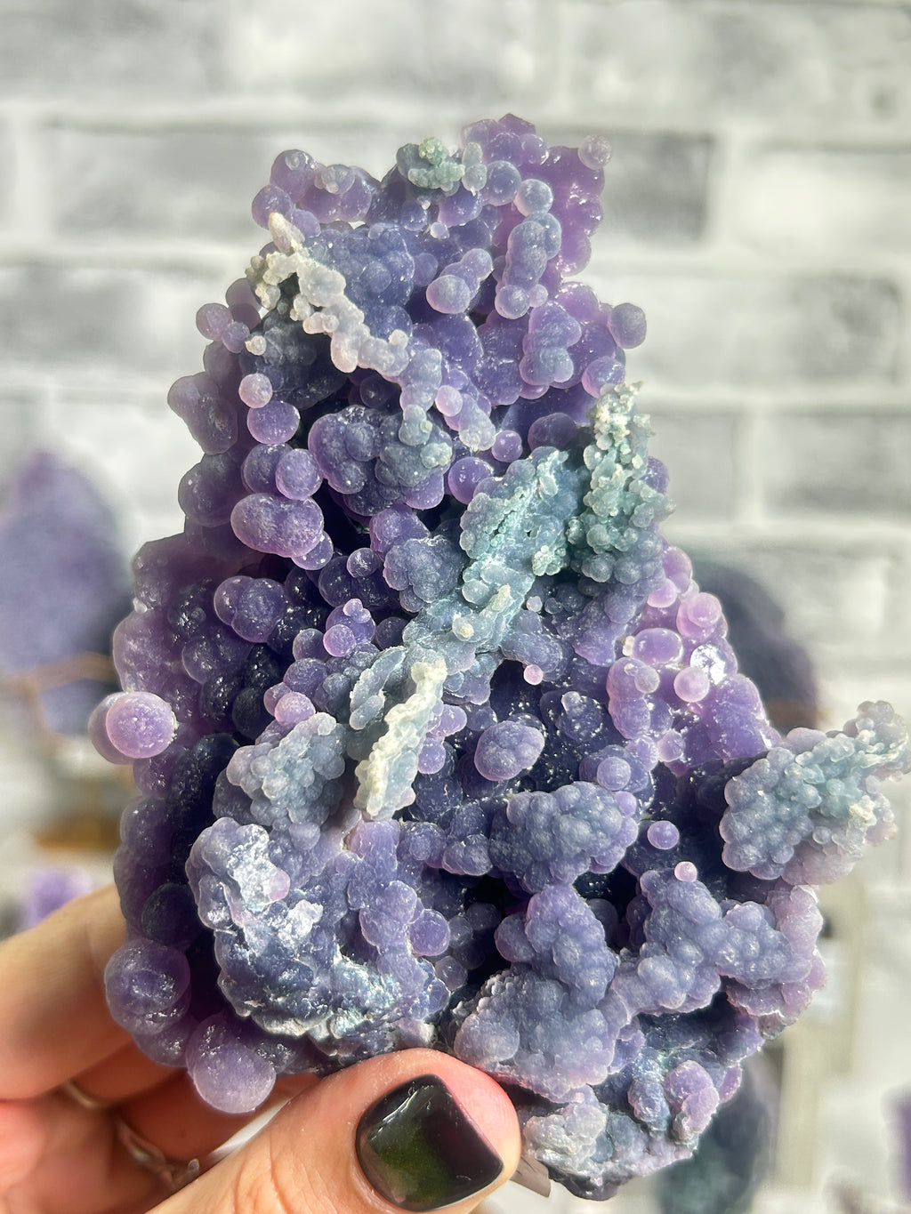 A hand with dark green nail polish holds a large, textured purple and blue mineral specimen with bubble-like formations against a blurred gray brick wall background.