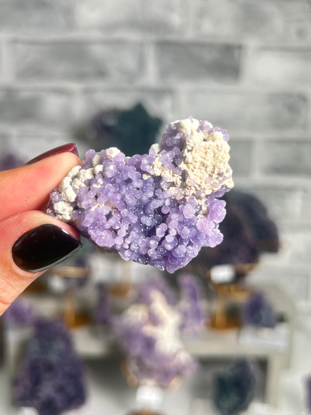 A hand with dark nail polish holds a purple amethyst crystal cluster with white calcite, set against a blurred background of other crystals and a gray brick wall.