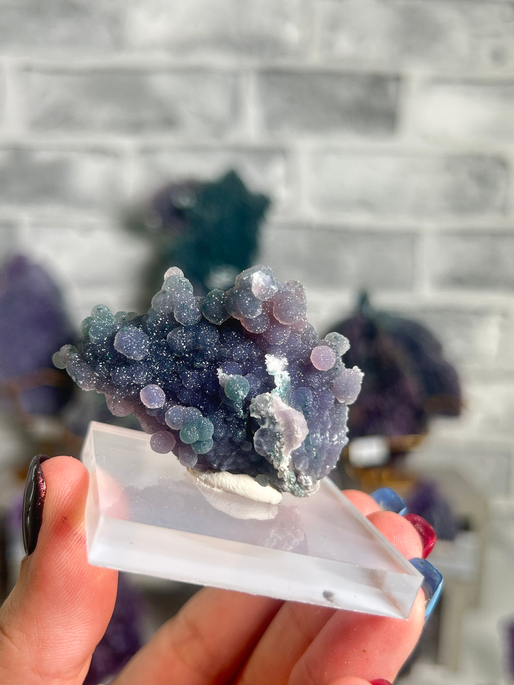 A hand holding a geode specimen of purple and blue druzy crystals on a clear acrylic display block, with a blurred gray brick wall background.