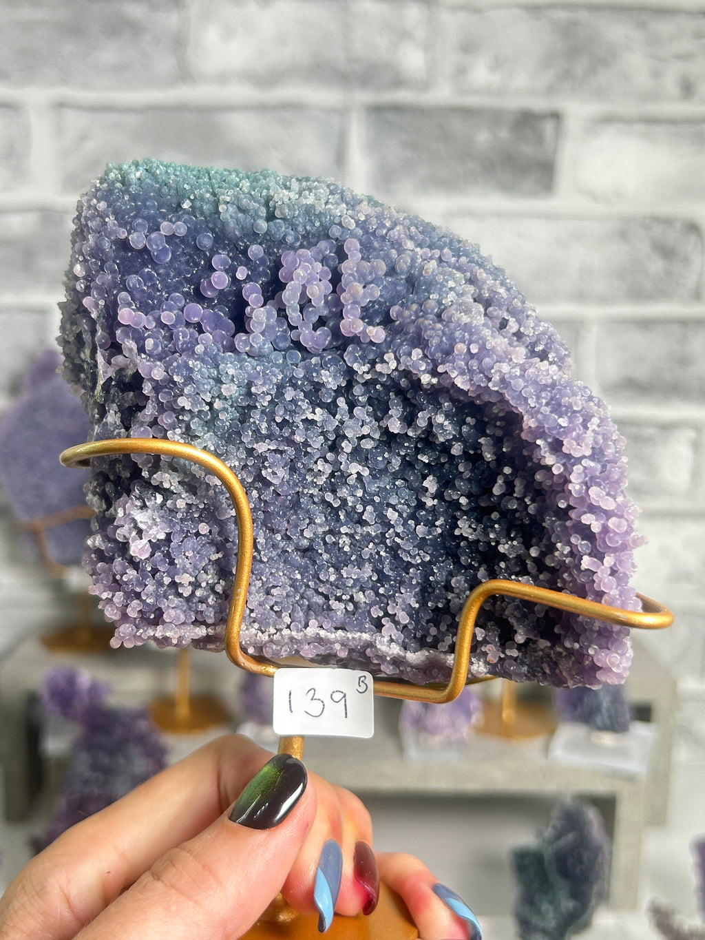 A purple and blue crystalline mineral specimen displayed on a gold wire stand, held by a hand with colorful nail polish in front of a gray brick wall background.