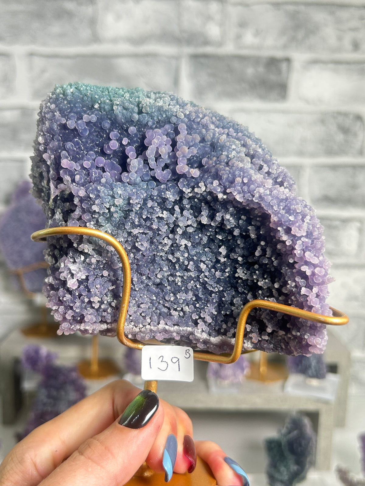 A purple and blue crystalline mineral specimen displayed on a gold wire stand, held by a hand with colorful nail polish in front of a gray brick wall background.