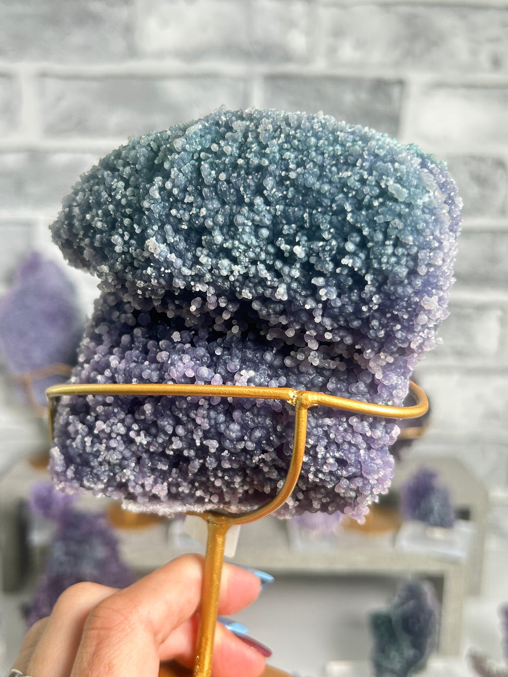 A close-up of a person's hand holding a purple and blue amethyst crystal cluster on a gold-colored metal stand, with a blurred gray brick wall background.