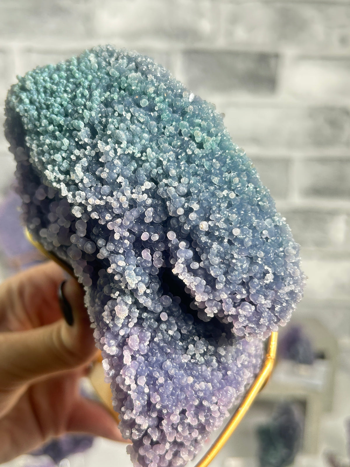 A close-up of a sparkling geode or crystal cluster with a blue-to-purple gradient, held by a hand against a blurred gray brick wall background.