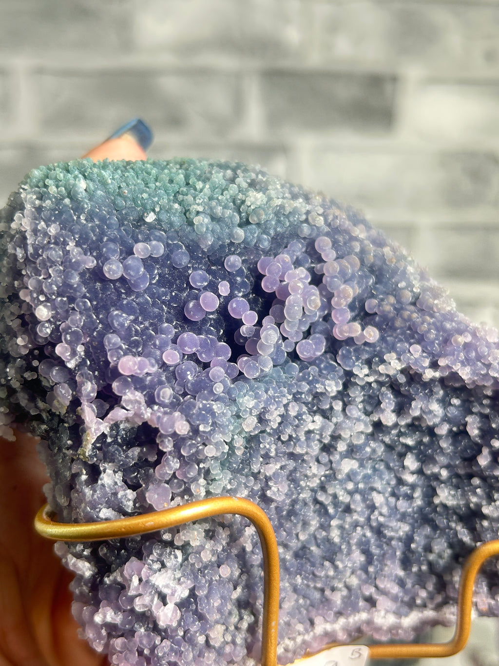 A close-up of a textured geode or crystal cluster with a blue-green to lavender-purple gradient, held in a gold wire display stand against a blurred gray brick wall background.