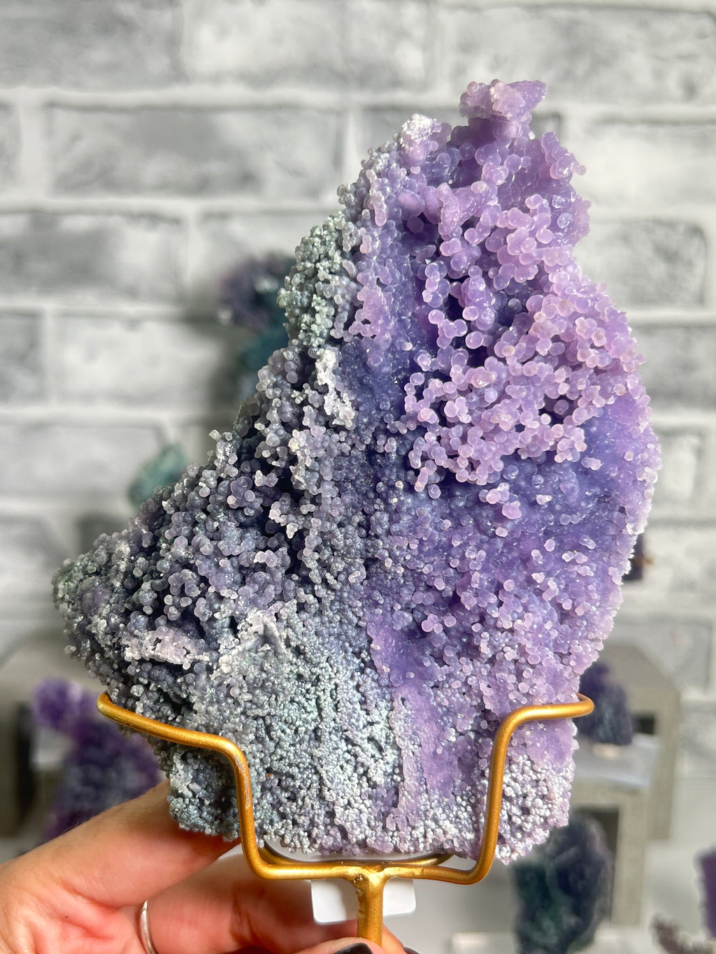A purple and lavender crystal geode specimen with textured, bubble-like mineral formations, displayed on a gold-colored metal stand held by a hand against a blurred gray brick wall background.