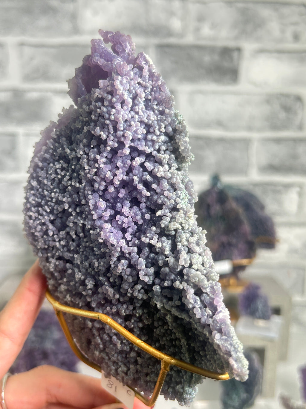 A purple and lavender amethyst geode specimen with a textured, crystalline surface, displayed on a gold-colored wire stand held by a hand against a gray brick wall background.