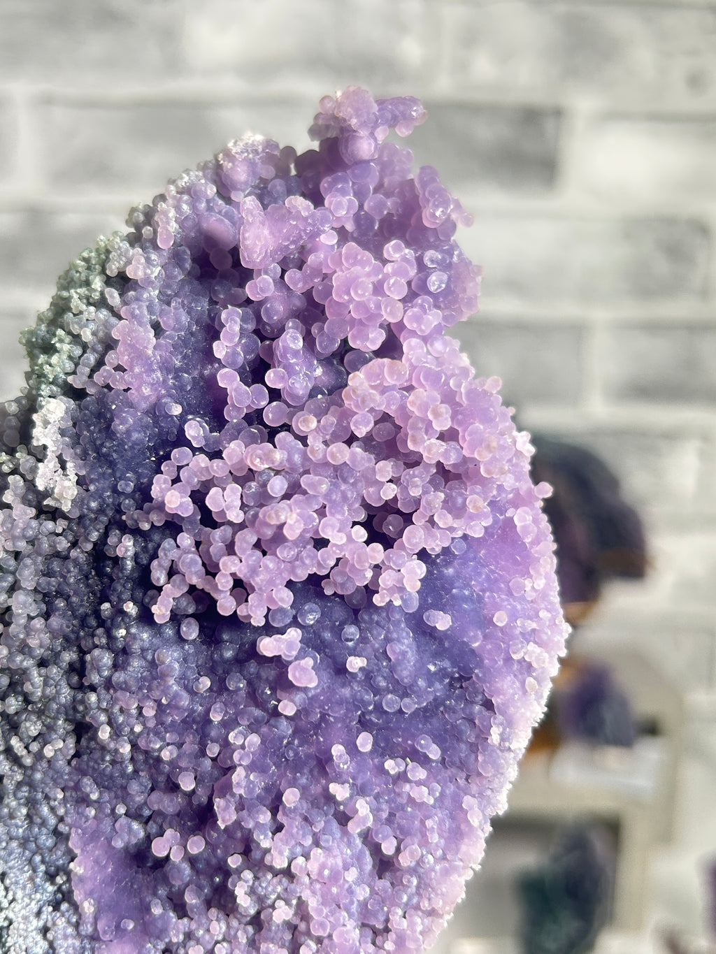 A close-up of a purple crystal geode specimen with dense, rounded lavender and lilac mineral clusters, set against a blurred gray stone background.